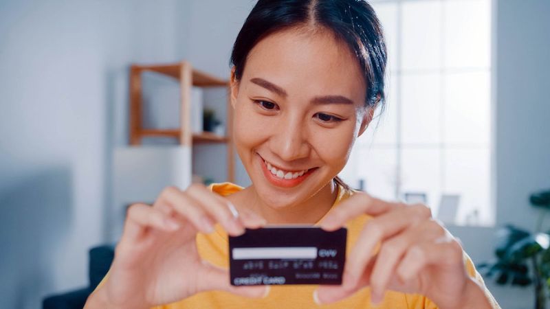 Young Asian Woman Holding Credit Card And Using Laptop Making Payment Online Sitting At Desk In Living Room At Home.